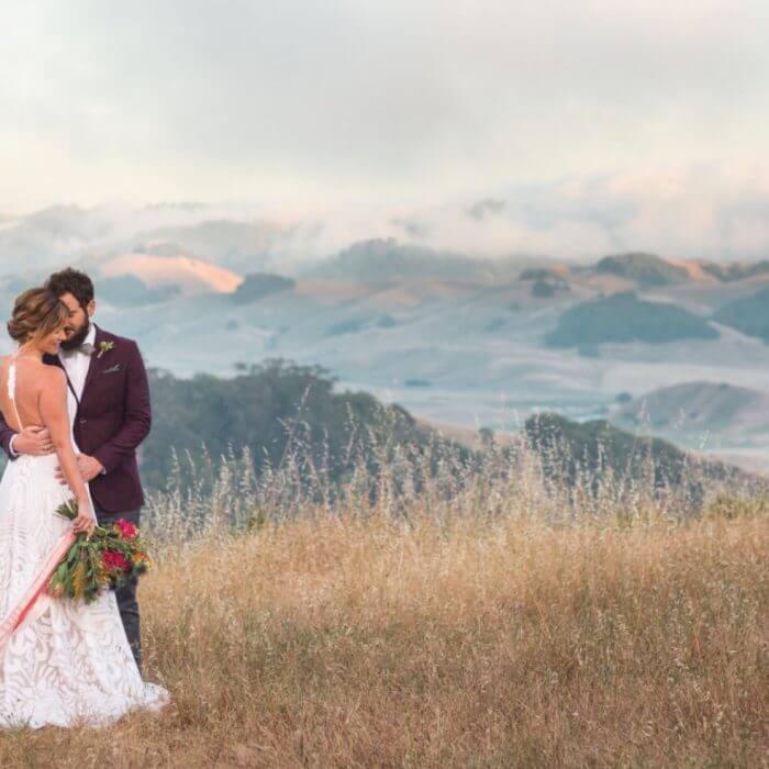 Groom embracing bride holding flower bouqet on top of a hill with hills and fog in the background, high end floral design by Aimée Lomeli Designs – Napa County florist – Sonoma County florist