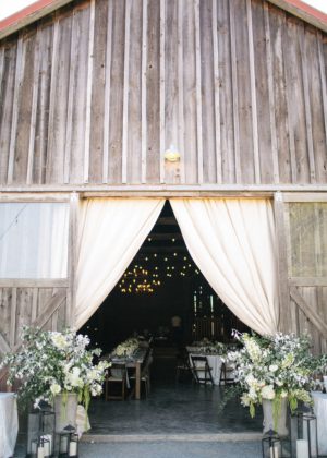 The front of a wooden barn, decorated with white curtains and floral arrangements, with tables set up inside for a wine country reception – floral design and styling by Aimée Lomeli Designs, Sonoma & Napa County florist