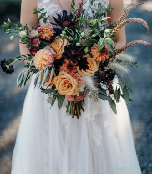 Bride in wedding dress holding bouqet of orange, pink, and green flowers in front of her; floral design by Aimee Lomeli Designs