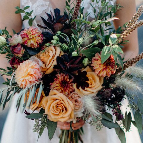 Bride in wedding dress holding bouqet of orange, pink, and green flowers in front of her – High-end floral design by Aimee Lomeli Designs