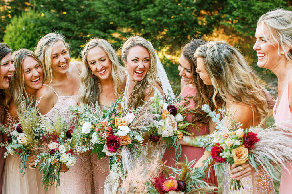 Group of 7 bridesmaids and bride holding wedding bouquets and laughing outdoors