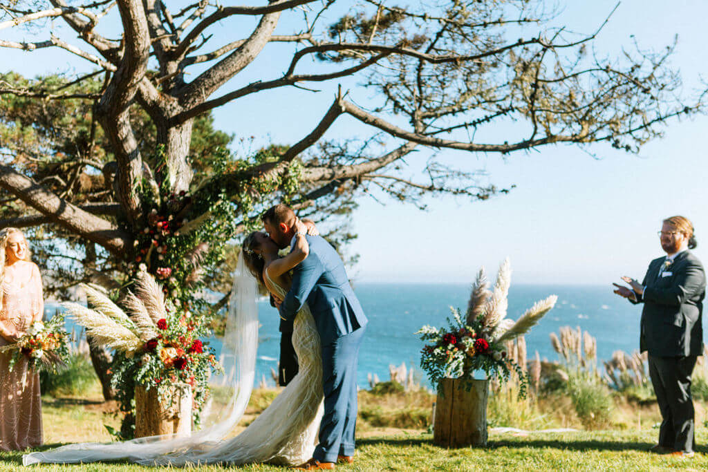 Bride in groom kissing in between floral decorated tree stumps, bridesmaid, and groomsmen, with Sonoma county ocean in background
