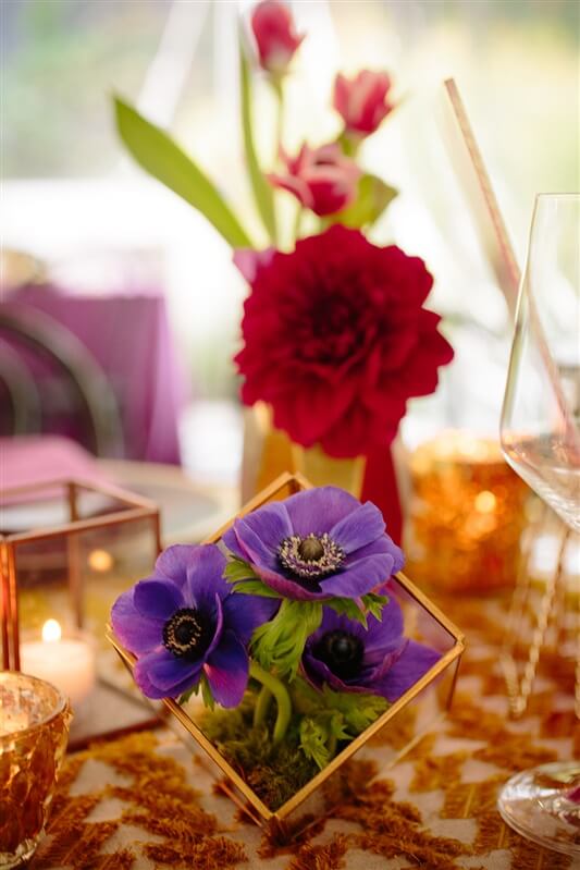 A purple floral arranegement set on table in front of a red floral arrangement and candles