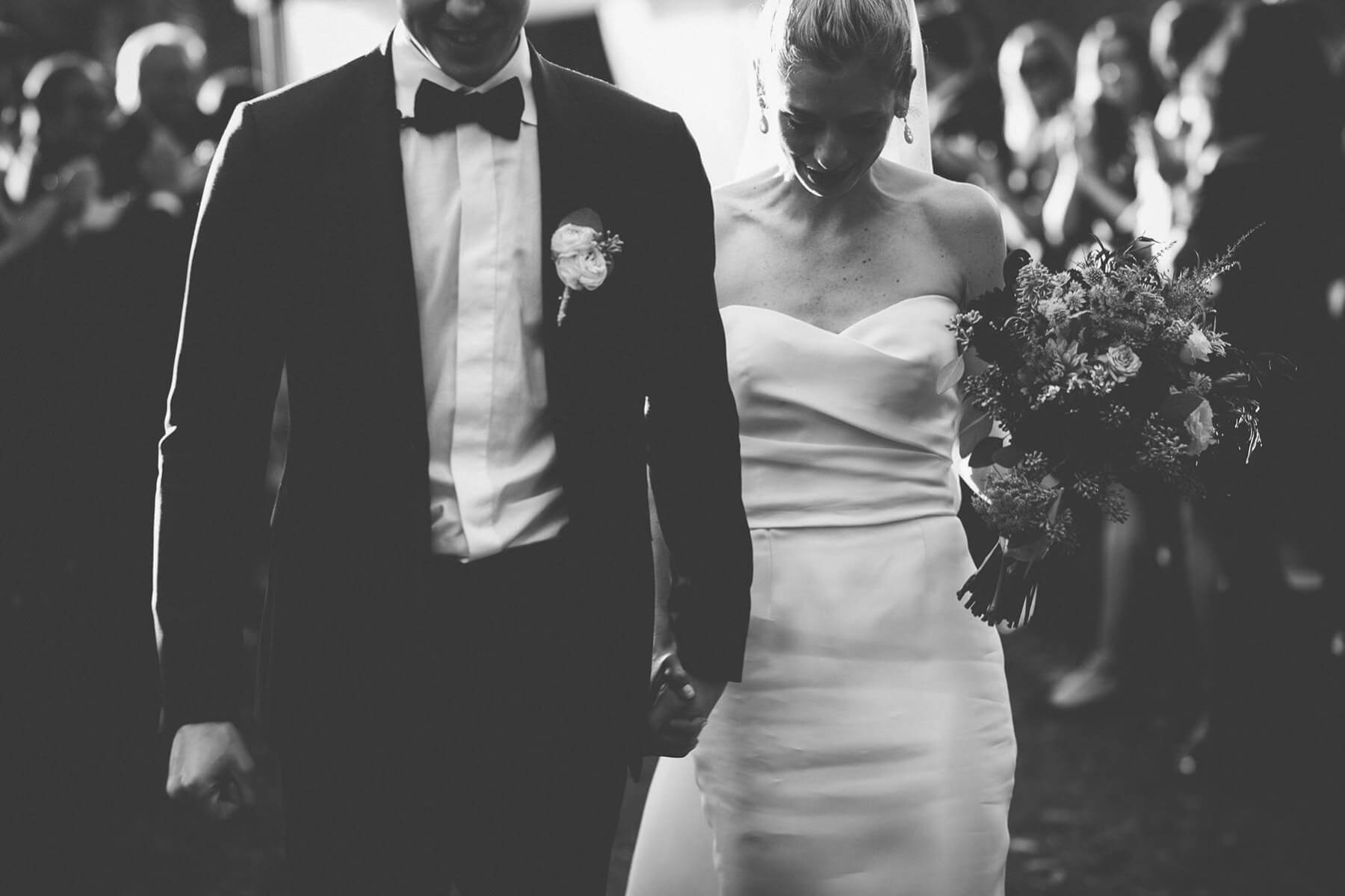 Black and white photo of newly married couple walking down aisle while bride holds wedding bouqet