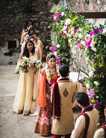 Bride and groom in front of floral wedding arch with bridesmaids holding bouqets in background at Kunde Ruins