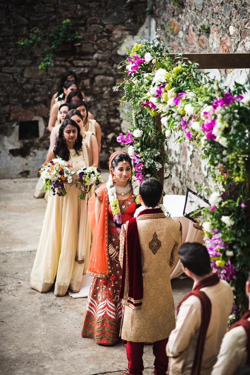 Bride and groom in front of floral wedding arch with bridesmaids holding bouqets in background at Kunde Ruins