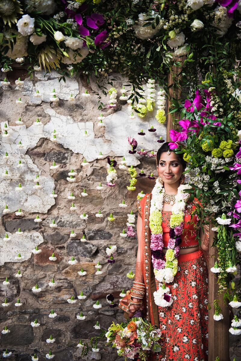 Bride holding bridal bouqet under floral wedding arch with rustic brick background in Kunde Ruins