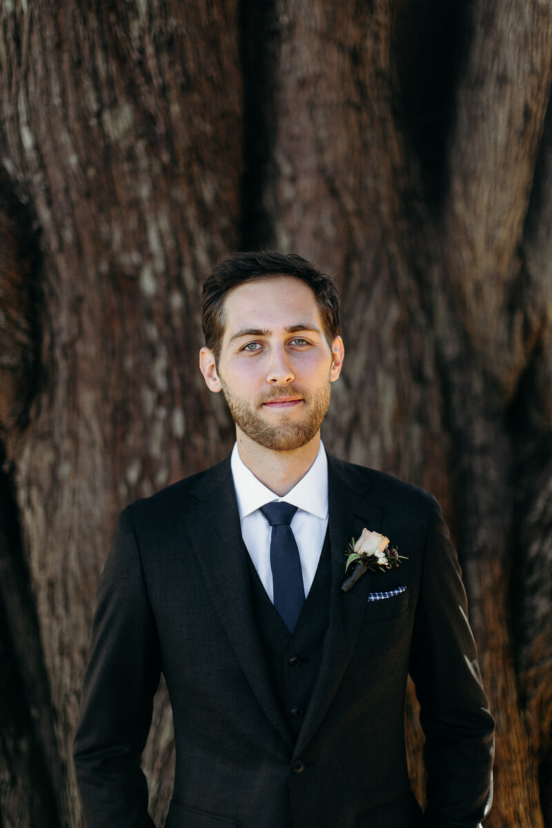 Groom with floral wedding corsage and tuxedo standing in front of tree