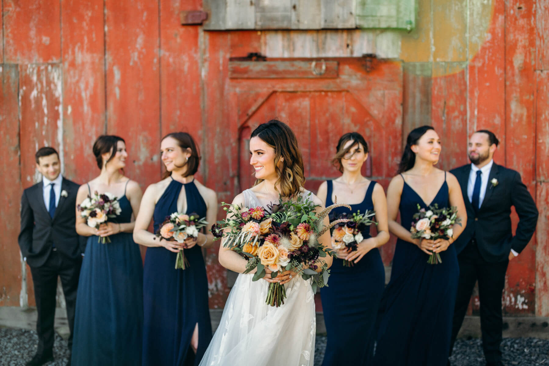 Bride and bridesmaids holding floral wedding bouquets with groomsmen and rustic barn in background at Stemple Creek Ranch in Marin County