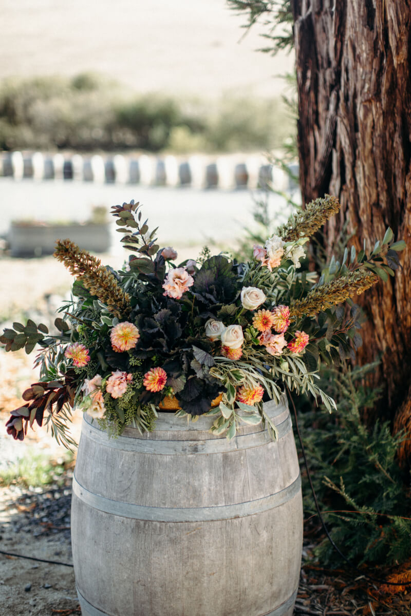 Rustic floral wedding arrangement set atop wine barrel outdoors in front of tree at Stemple Creek Ranch
