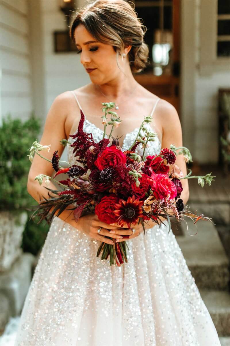 a bride outdoors in a white sparkly dress holding an extravagant bouquet of red and maroon flowers