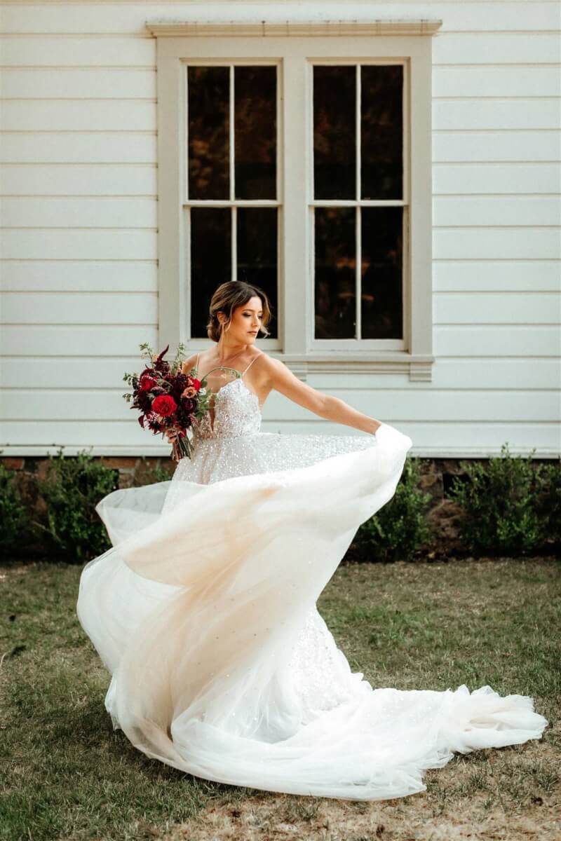a bride holding a red bouquet in front of a white building and window holding her skirt out while she turns