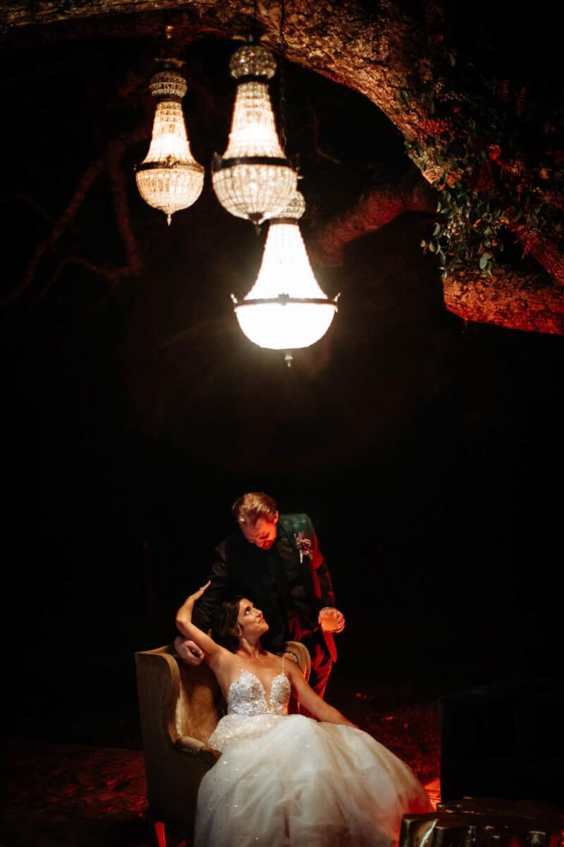 a bride sitting in a chair outdoors at night looking up at a groom while lights hang from a tree above