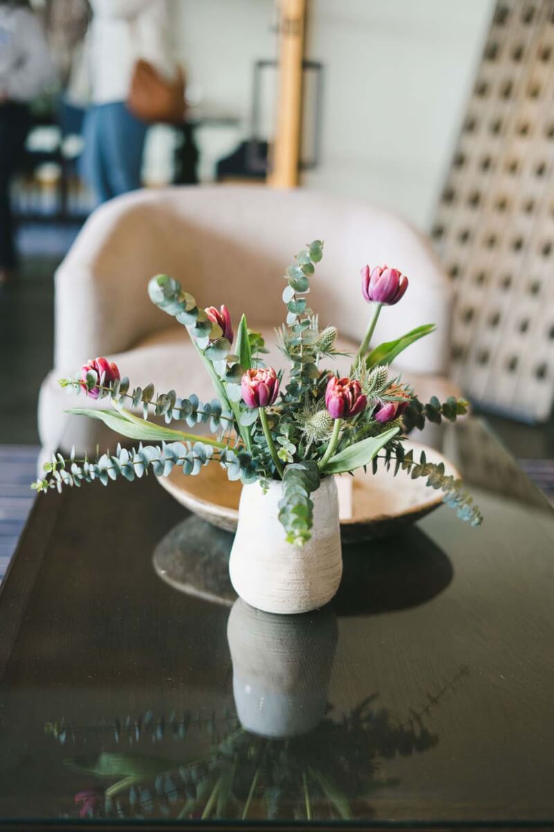 elegant pink flowers and greenery in a natural white vase on a wooden table with blurred people in the background