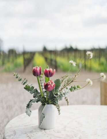 an elegant arrangement of pink and white flowers and greenery in a white vase on an outdoor vineyard table