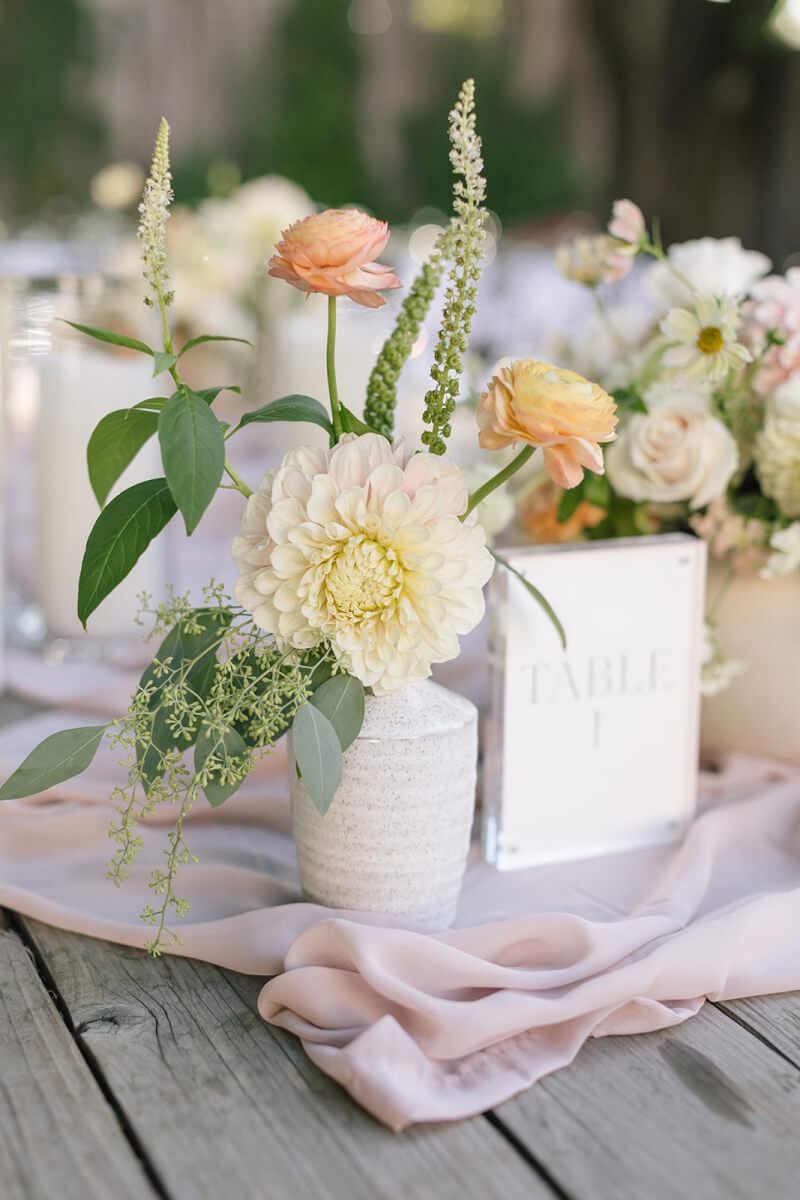 a wedding table topped with a vase filled with soft peach and buttercream flowers.