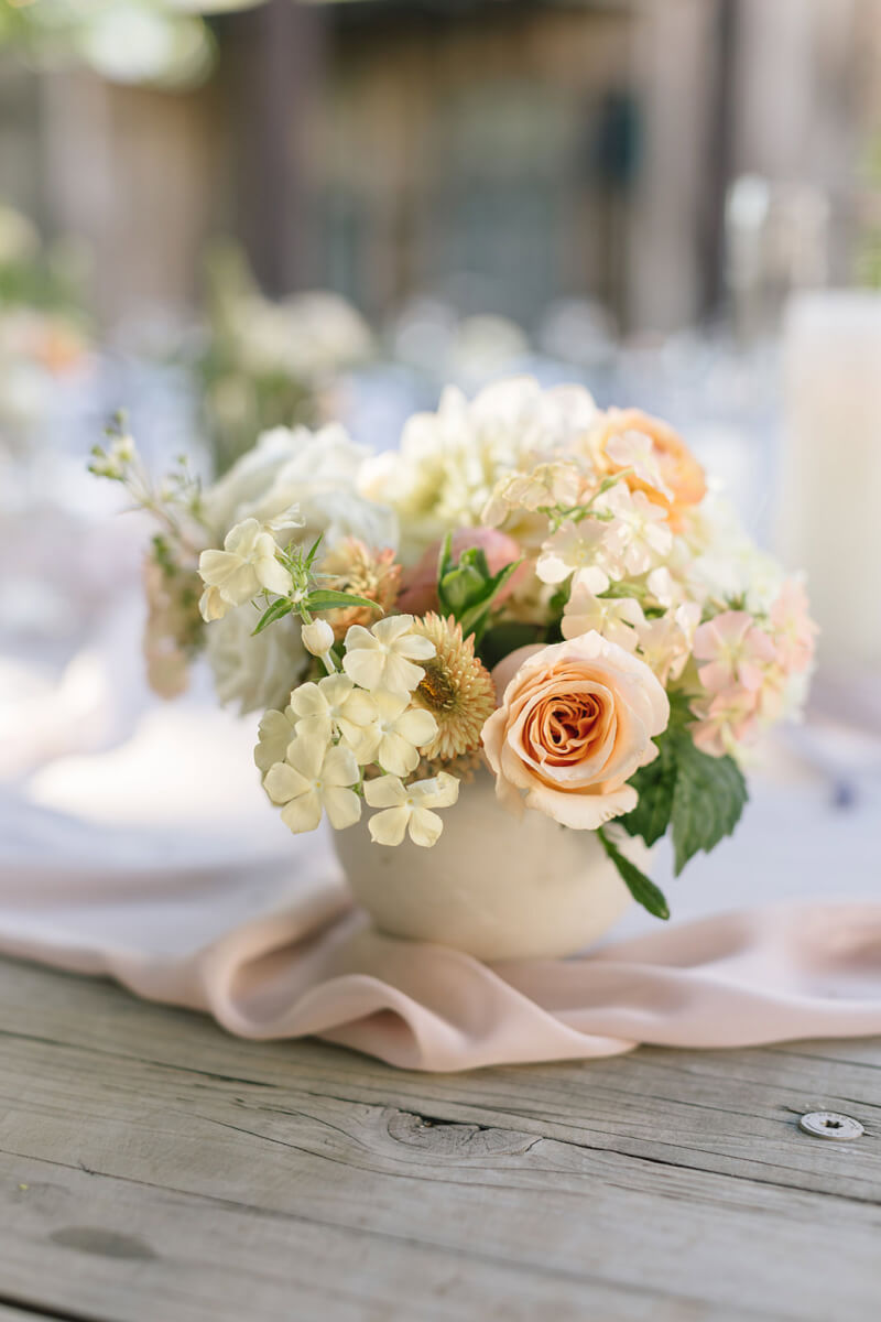 a white vase filled with flowers by Aimee Lomeli Designs on top of a wooden table.