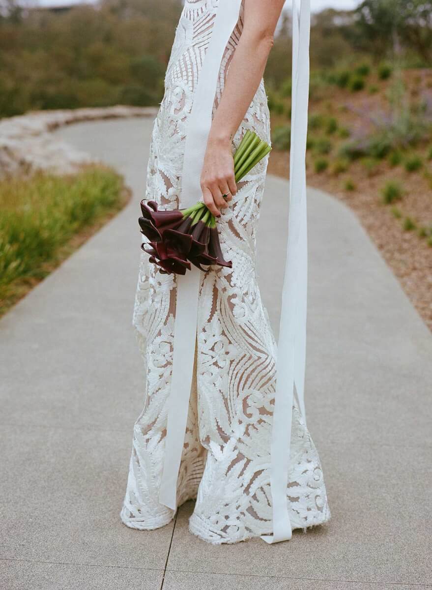 close up of the bottom of the bride's lace wedding dress, her hand holding a bouquet of plum calla lilies
