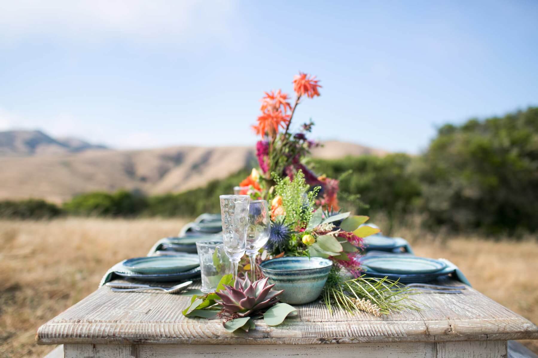 a wooden table topped with plates and vases filled with colorful flowers for an outdoor wine country private ranch event