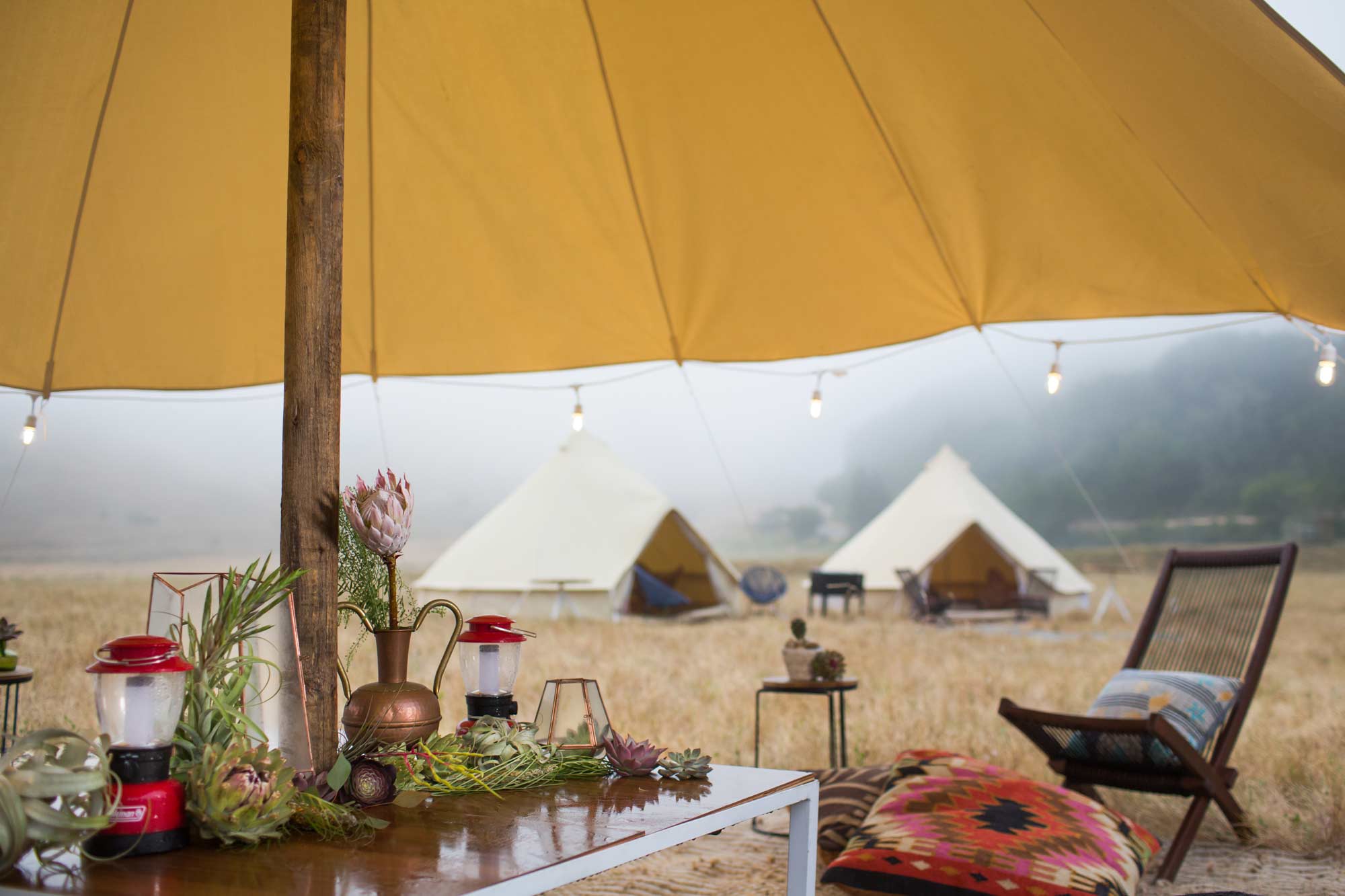 a table with florals and camp lanterns under a yellow umbrella with tents in the background at a wine country retreat