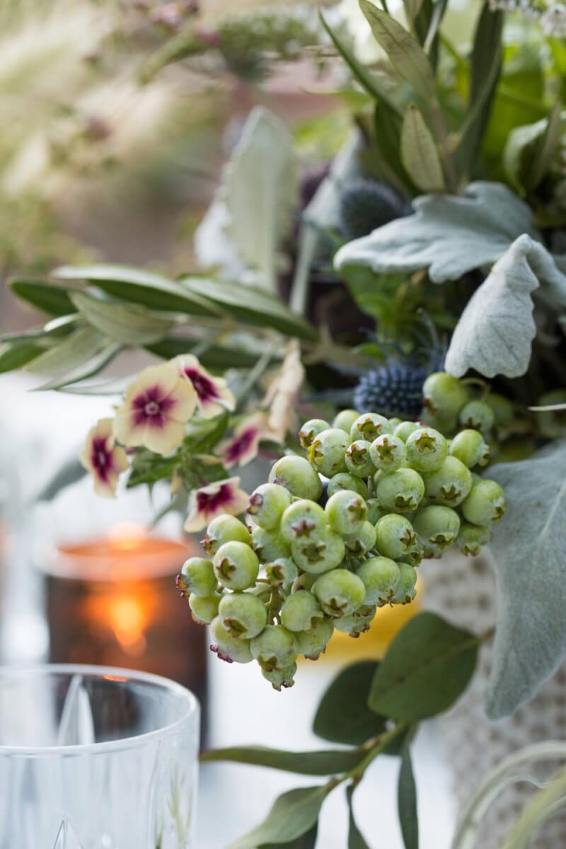 a close up of a vase with flowers and green berries on a table set for a private garden party