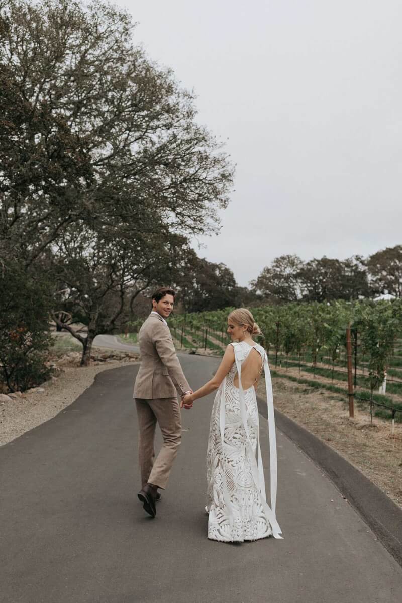 the bride and groom walking down the winery road holding hands.