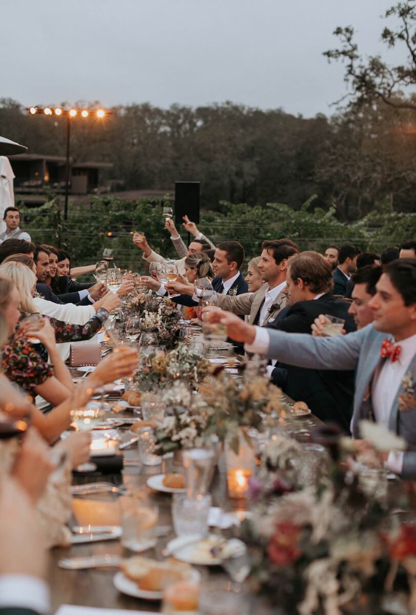 wedding guests raising their glasses sitting at a long table adorned with flowers