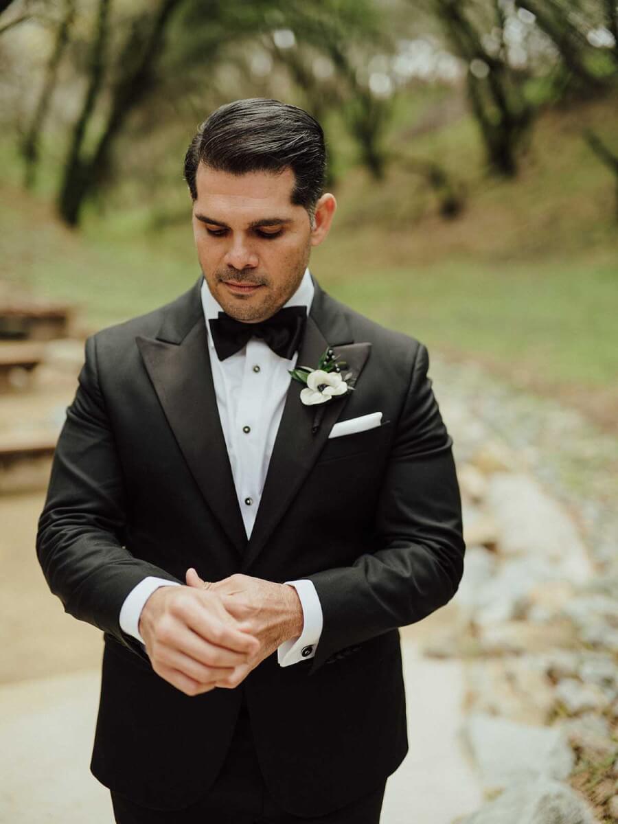 A groom in a black tuxedo standing in a wooded area.