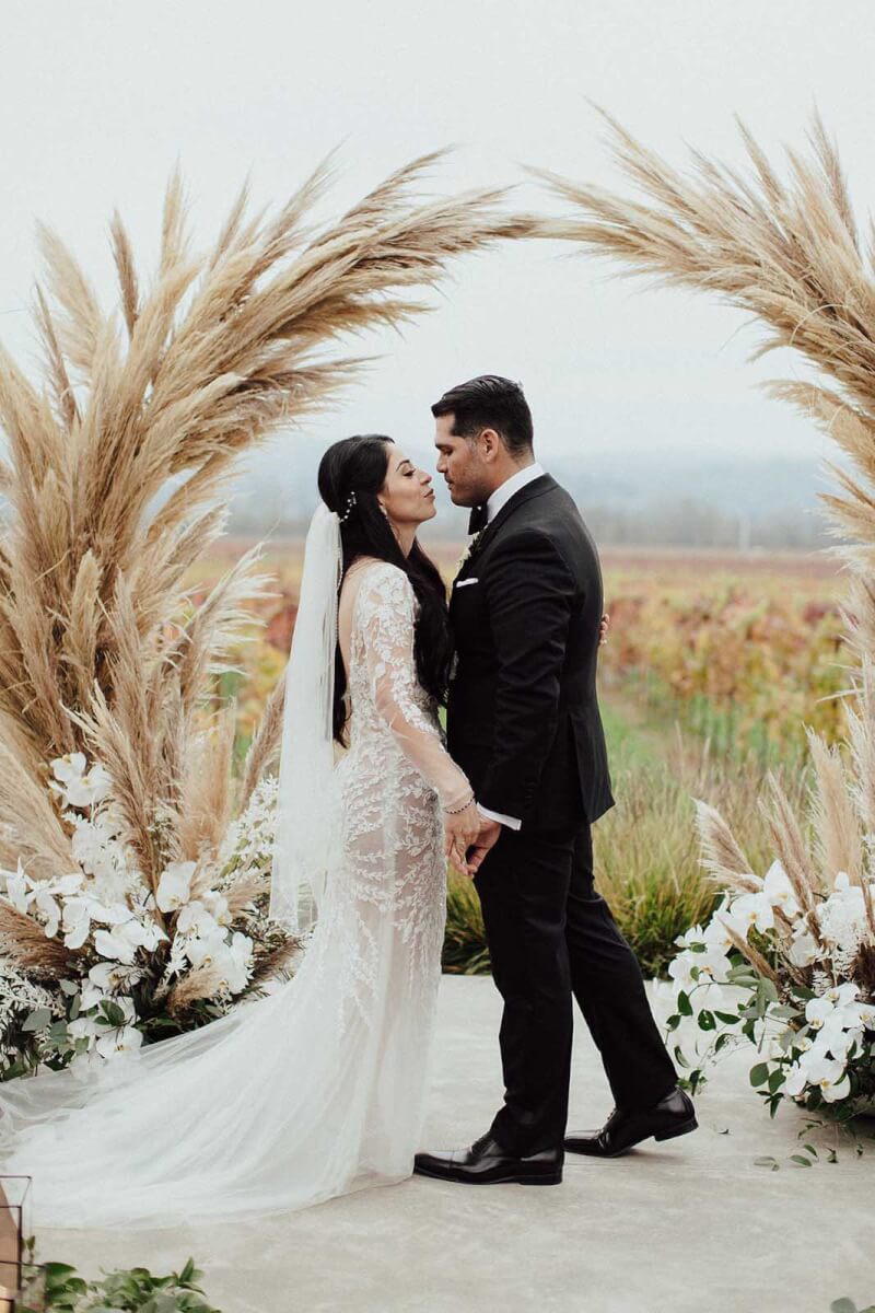 the bride and groom under the pampas grass wedding arch