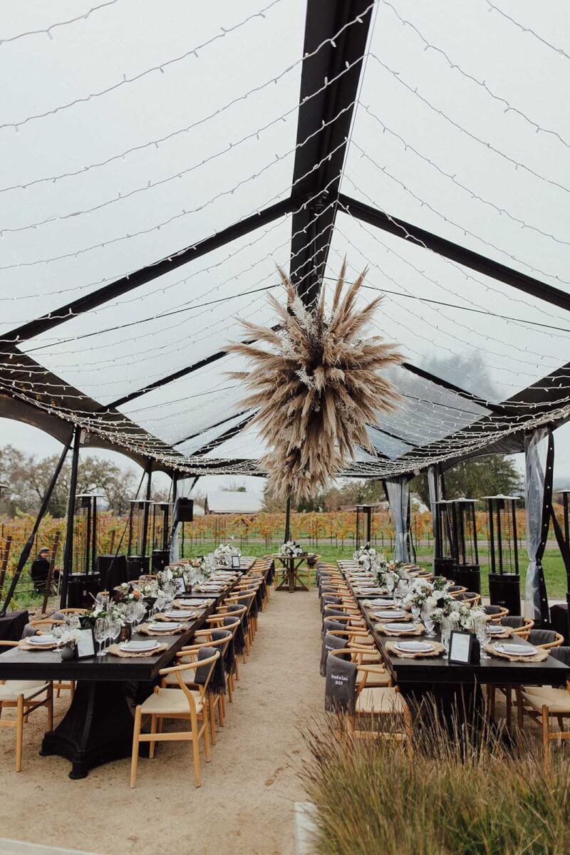 the wedding tables under a canopy of lights in the vineyard with black and white bouquets and pampas grass chandelier style arrangements