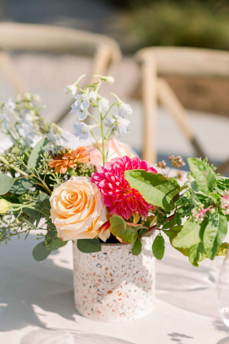 A wedding table bouquet in a cylindrical vase with blue, bright pink and coral flowers and greenery.