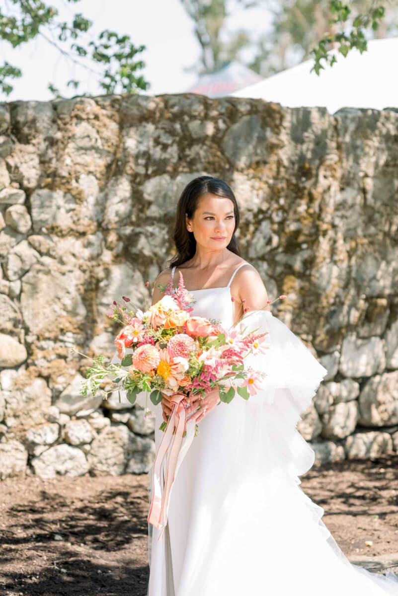 The bride in front of a stone wall holding the wedding bouquet in playful colors of coral, pink, and orange with pink ribbon.