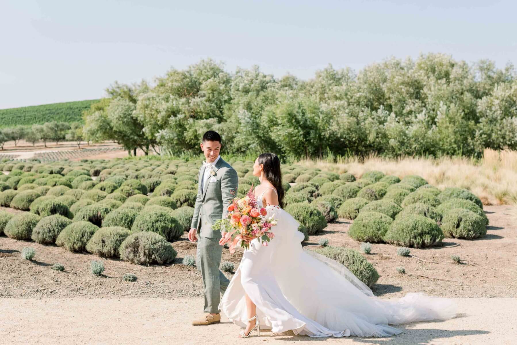 The groom and bride, holding the bridal bouquet of colorful butterfly ranunculus, cosmos, and dahlias, walk through a vineyard.