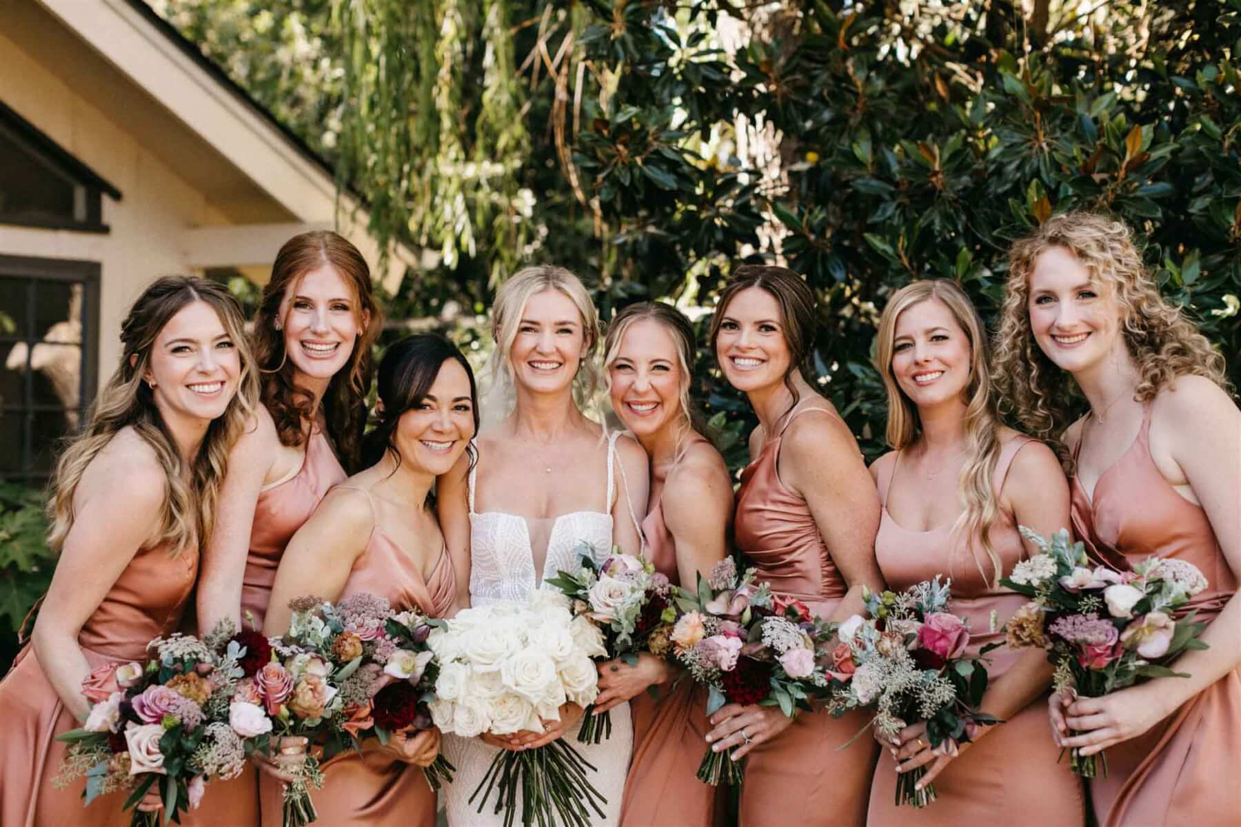 The bride holding an all-white bridal bouquet, and seven bridesmaids in rose-colored dresses holding bouquets in shades of deep plum, coral, peach, sage, and forest green.