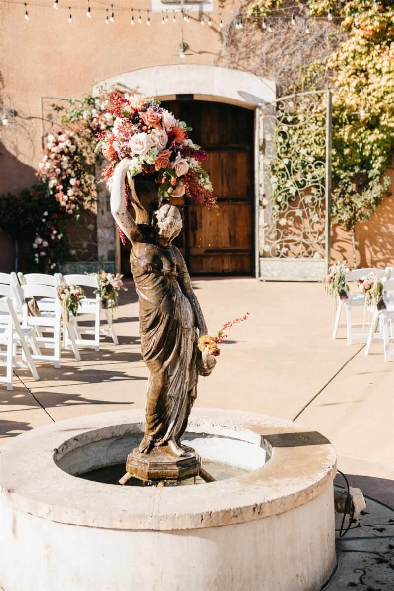 Elegant wedding blooms top an Italian statue fountain, with more blooms in the background on the rod iron gates.