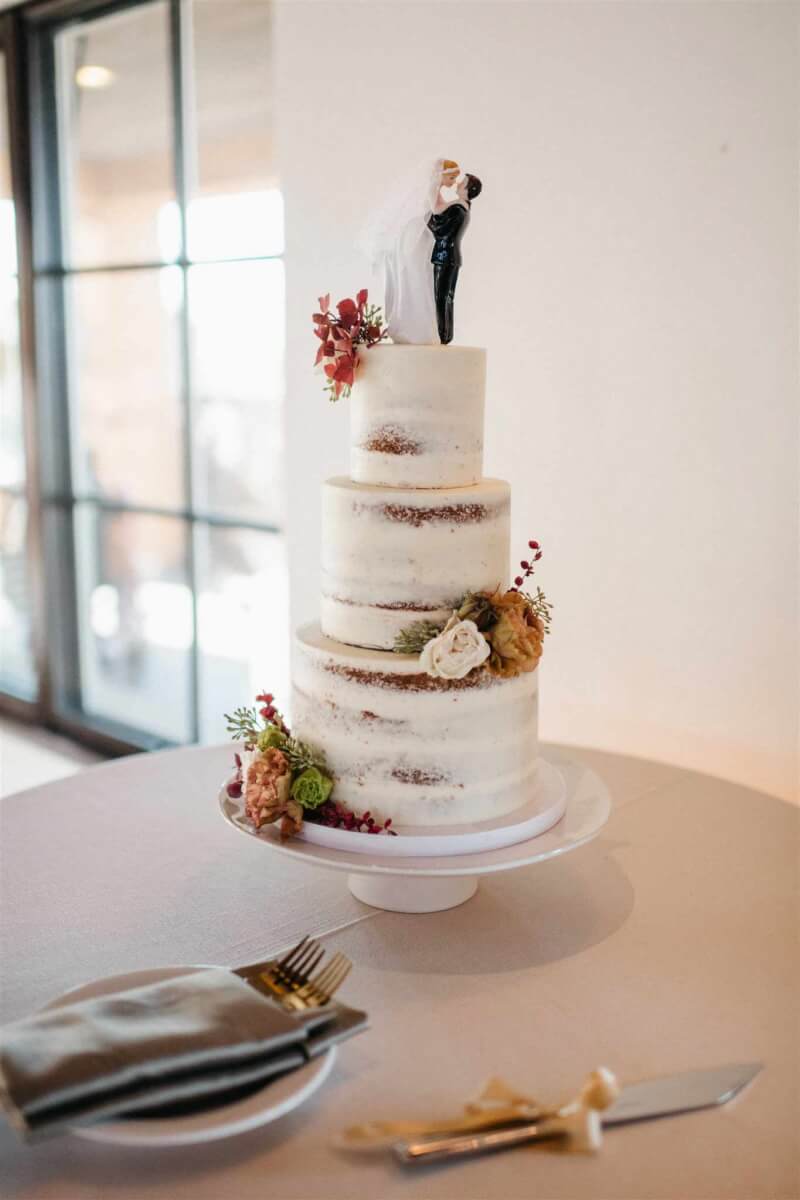 The tiered wedding cake with naked frosting style, elegant floral accents, and a bride-and-groom cake topper.