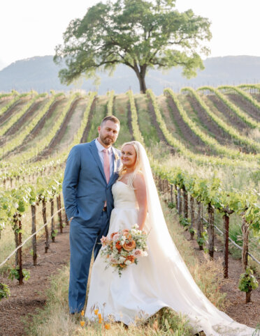 A bride and groom stand in a vineyard, with rows of grapevines and a large tree in the background. The bride holds a bouquet and wears a veil.