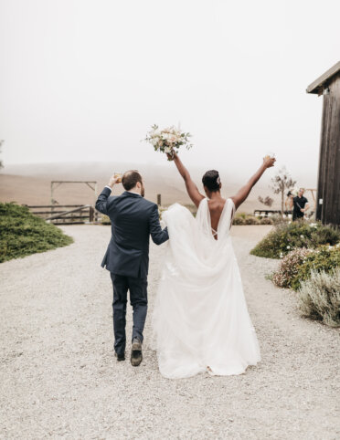 A bride and groom walk down a gravel path, with the bride holding a bouquet and raising her arms in celebration. The scene is outdoors with rustic wooden structures nearby.