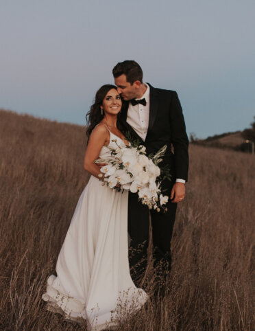 A bride in a white gown holds a bouquet of white flowers, standing beside a groom in a black tuxedo, in a grassy field.