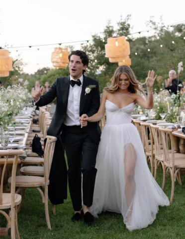 The bride and groom walk hand in hand outdoors beside the long banquet tables, smiling and waving, surrounded by Aimee Lomeli Designs table flower arrangements and string lights.