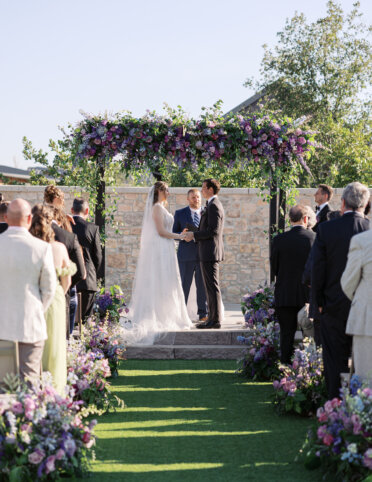 The bride and groom stand under the flower-covered arch, holding hands during the outdoor wedding ceremony, with guests seated on both sides of the aisle.